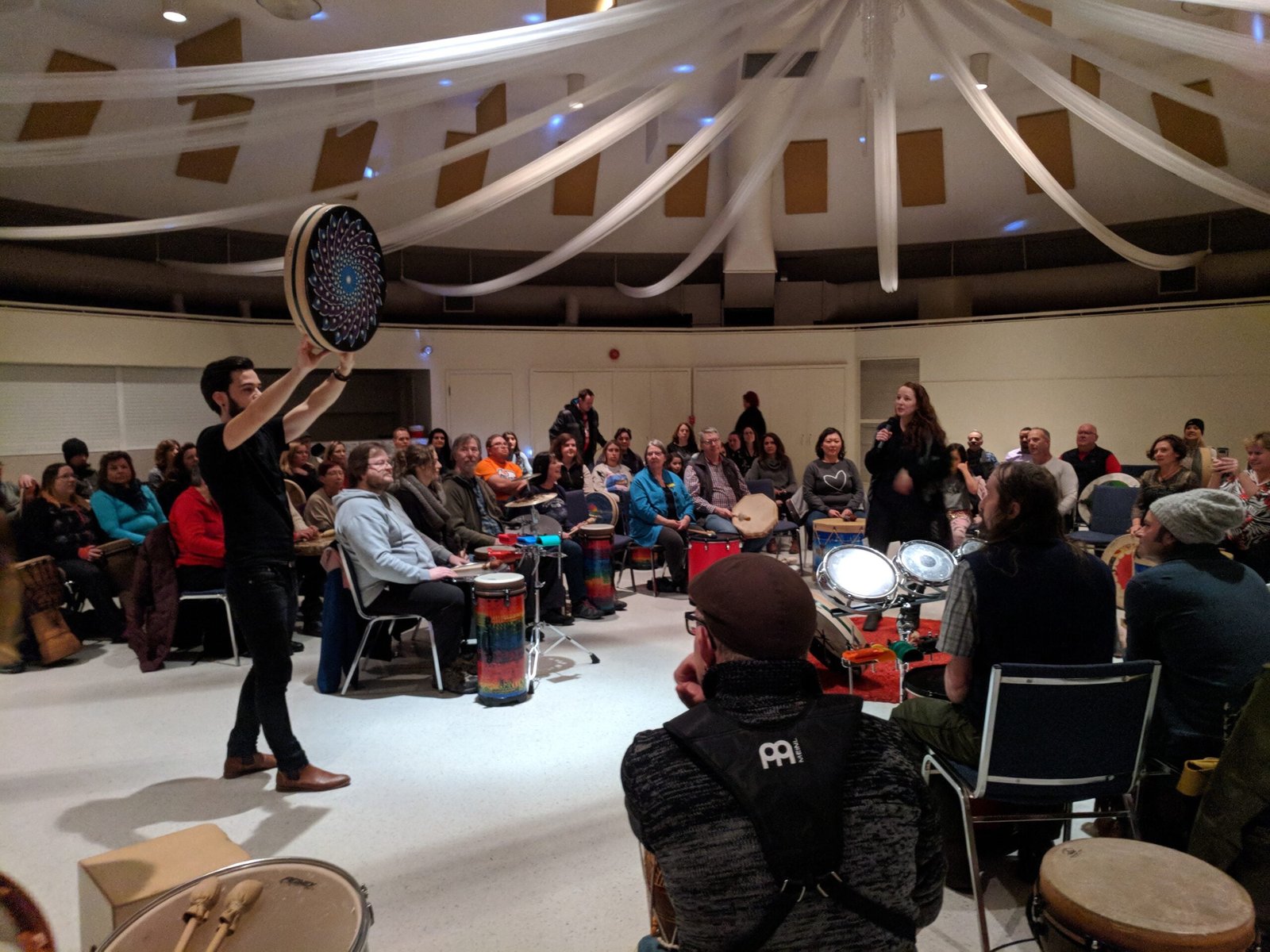 A Circles of Rhythm facilitator holds up a hand drum while leading a full room of seated participants, each with their own drum