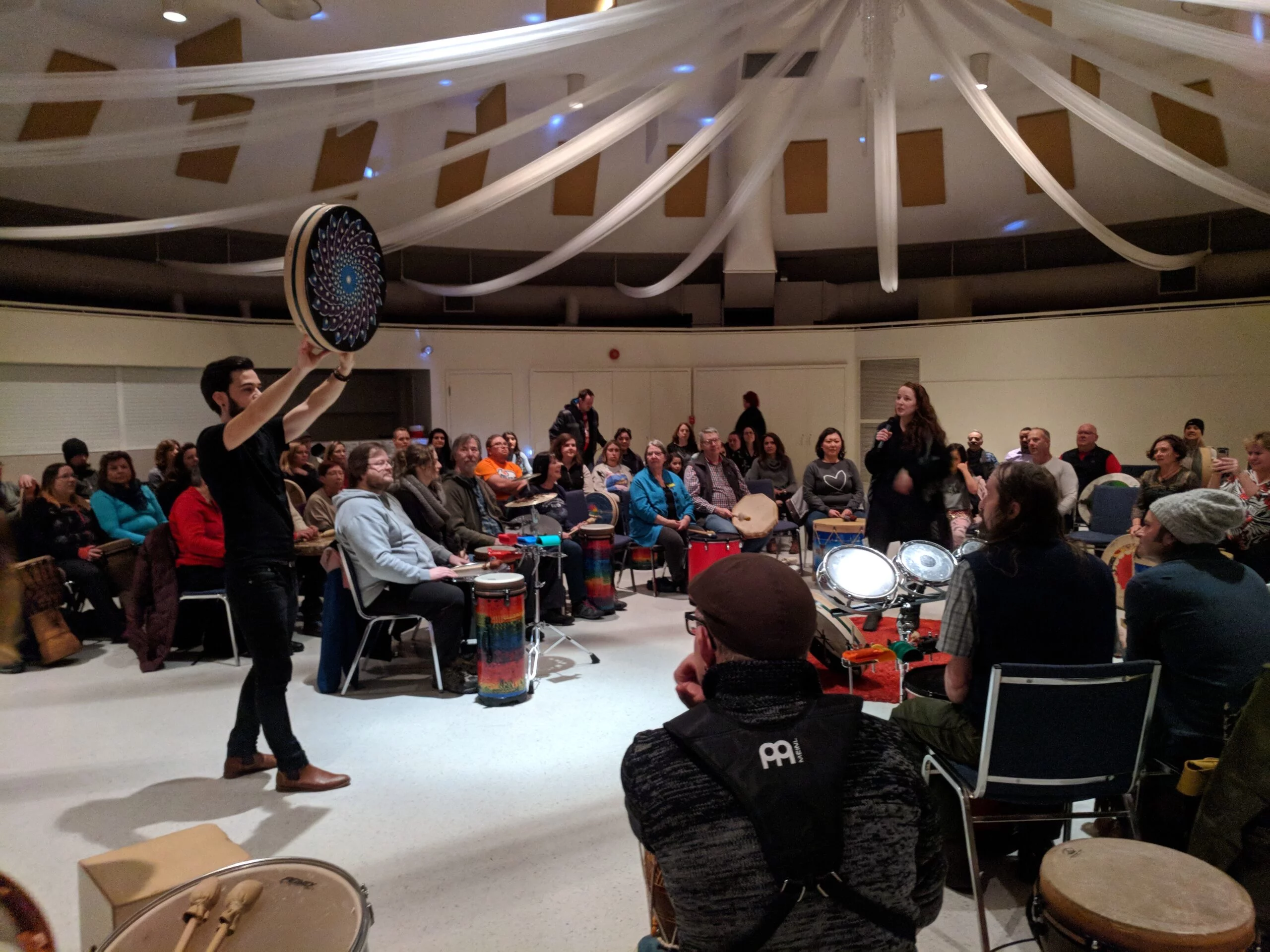 A Circles of Rhythm facilitator holds up a hand drum while leading a full room of seated participants, each with their own drum